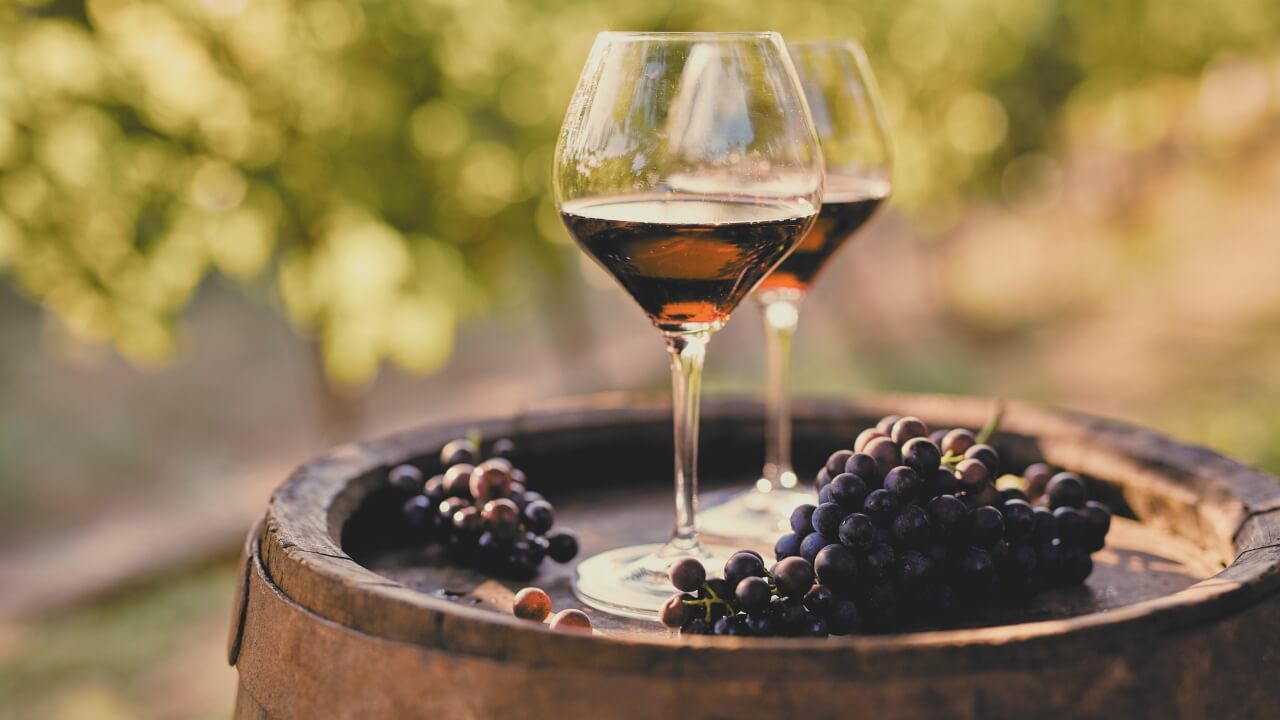 Duas taças com vinho tinto em cima de um barril de vinho feito de madeira com uvas. Ao fundo uma plantaçõa de uva.