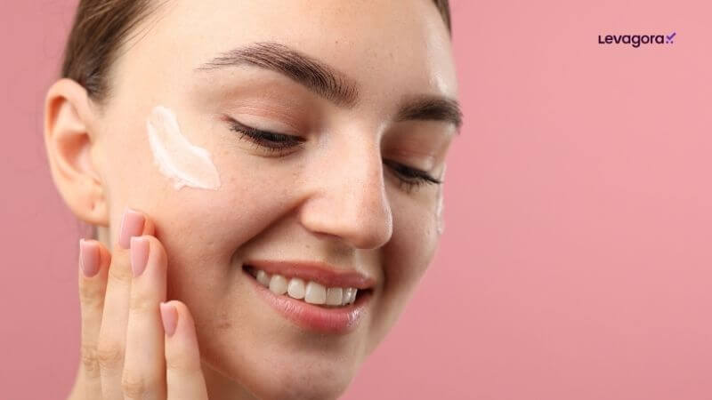O Segredo do Skincare Foto em close de uma mulher com a pele limpa e olhos fechados, sorrindo gentilmente sobre um fundo rosa. Ela tem um toque de creme branco na bochecha e toca o rosto com a mão, que tem unhas pintadas de rosa claro e dois anéis dourados. No canto superior direito, há um pequeno logotipo roxo escrito "Levagora".