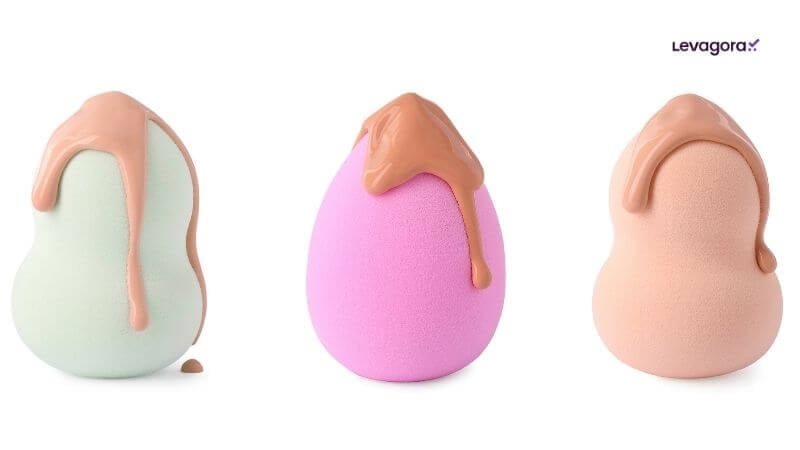 Acessórios de Maquiagem e Economia Três esponjas de maquiagem dispostas em uma linha contra um fundo branco puro. Cada esponja tem base de maquiagem líquida gotejando pelo topo e pela lateral. Da esquerda para a direita: uma esponja verde-clara com maquiagem bege-clara; uma esponja rosa-choque com maquiagem bege-escura; e uma esponja bege com maquiagem bege. O logotipo "Levagora:" está no canto superior direito.