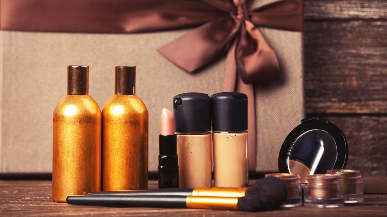 Como montar kits de beleza - Uma variedade de produtos de maquiagem, incluindo dois frascos dourados, bases, batom rosa claro e pincéis, estão arrumados em uma superfície de madeira marrom escura. Ao fundo, vê-se uma caixa de presente dourada com um laço marrom.