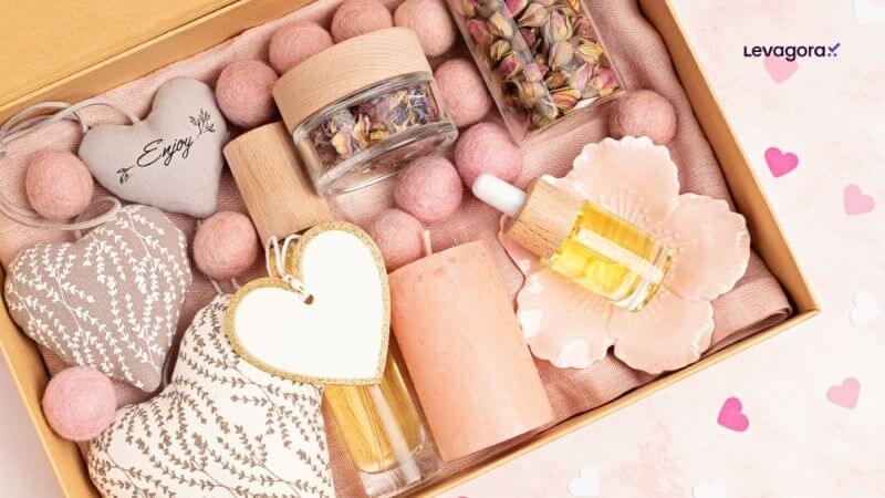 kits para o calendário comercial Uma caixa de presente aberta, feita de papel kraft, revela uma seleção de itens de autocuidado em tons de rosa e lavanda. Dentro, encontram-se travesseiros decorativos em forma de coração com o texto "Enjoy", sabonetes e velas com aroma de rosas, pétalas secas, óleo essencial e decorações esféricas felpudas cor-de-rosa. O fundo é rosa claro com corações decorativos e o logotipo da Levagora.
