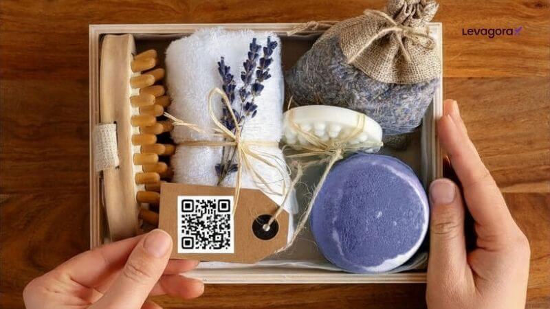 Kits de Autocuidado e SPA Uma mão segura uma caixa de madeira aberta, preenchida com itens de spa de lavanda e autocuidado. A caixa inclui uma escova corporal com cerdas de madeira, uma toalha branca enrolada e amarrada com raminhos de lavanda seca, uma barra de sabonete de spa, um sabonete circular roxo e uma bolsa de pano cheia de lavanda seca. Uma etiqueta de papel kraft pendurada em uma corda exibe um código QR, e o logotipo da Levagora está no canto superior direito.