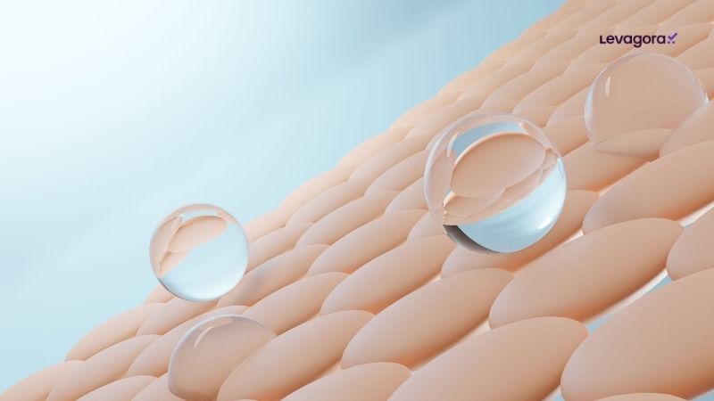 Invista em skincare Representação digital em 3D de uma superfície de pele em close-up, composta por células ovais e organizadas. Sobre essa superfície, gotículas de água cristalinas e esféricas repousam, sugerindo hidratação profunda. O fundo é azul claro e o logotipo "Levagora" aparece no canto superior direito.