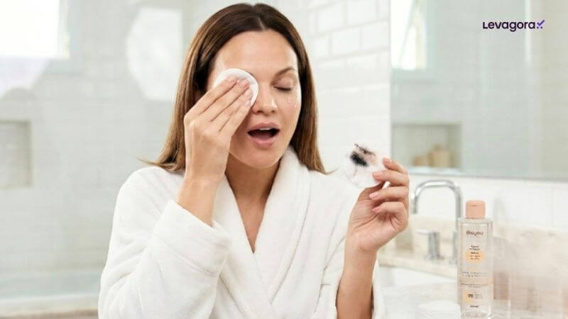 Dica como usar Água Micelar Detox Bisyou Uma mulher de roupão branco remove a maquiagem dos olhos com um disco de algodão em um banheiro. Ela está com o olho direito fechado enquanto aplica o algodão, e sua boca está ligeiramente aberta em uma expressão de concentração. Na mão esquerda, ela segura um segundo disco de algodão que já mostra resíduos de maquiagem preta. A garrafa de Água Micelar Bisyou está sobre o balcão à direita. O logotipo "Levagora✓" está no canto superior direito.